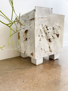 white textured ceramic sculpture resembling a footed cube by caroline blackburn sitting on a cement floor next to a tall plant