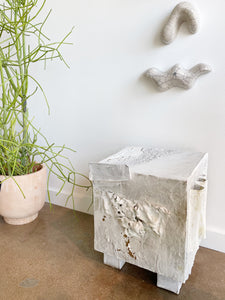 white textured ceramic sculpture resembling a footed cube by caroline blackburn sitting on a cement floor next to a tall plant